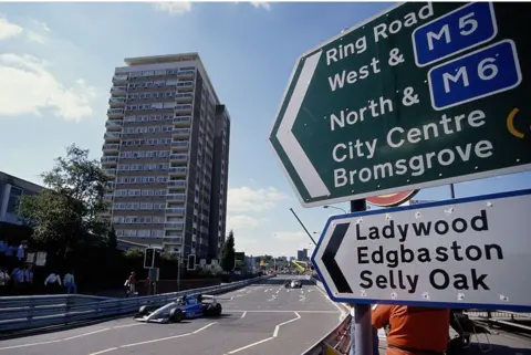 Getty Images A Formula 3000 racing car can be seen driving on Birmingham's middle ring road with some crowds looking on. In the background a tall block of flats can be seen next to the road. In the foreground road signs signal directions to Ring Road West & M5, North & M6, City Centre, Bromsgrove, Ladywood, Edgbaston and Selly Oak. 
