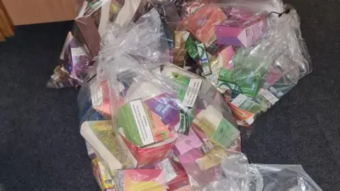 Lincolnshire Police A large quantity of colourful vapes products wrapped in plastic bags laid out on a dark-coloured carpet 