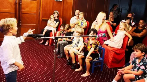 A boy is singing into a microphone as children and adults watch from chairs.