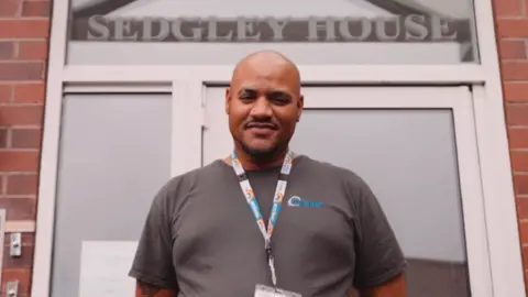 A mid-shot of a bald man standing in front of a doorway with a sign above it that reads "Sedgley House". He is wearing a grey T-shirt and has his hands behind his back. He also has a lanyard with rainbows on it around his neck. 