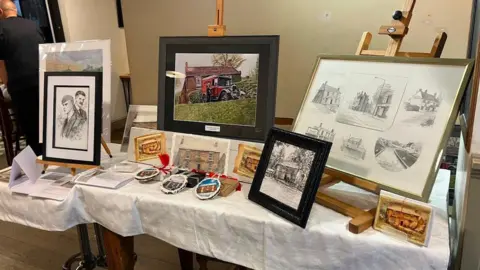 A table with a white table cloth on it is loaded with photographs and small ornaments depicting the former Crooked House pub in its fully-built state. 