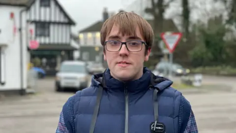 A young man with long light brown hair. He wears navy glasses and wears a navy puffer gilet with a navy and red checked shirt underneath. He is standing next to a road.
