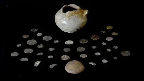 The pot, its lid and the coins are laid out on black fabric. The coins are of different sizes.