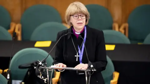 Jordan Pettitt/PA Wire A woman priest wearing black cassock dog collar and cross speaking at a lectern