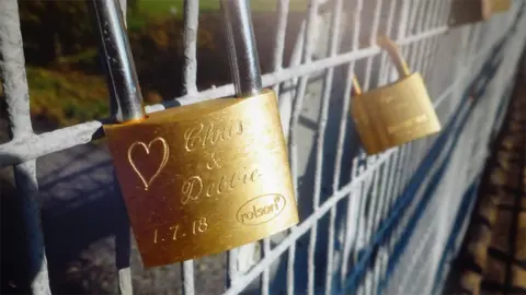 A gold padlock on a bridge with a heart engraved and writing saying Chris and Debbie 1.7.18