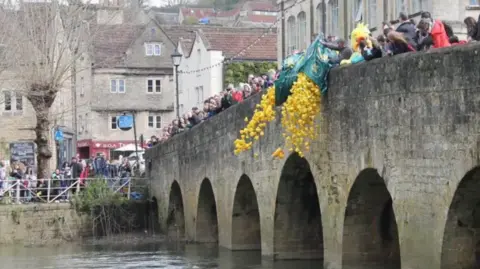 Bradford-on-Avon Town Council Crowds of people stand on an old bridge in Bradford-on-Avon as bags of rubber ducks are thrown into the water. Near the bridge, people stand and watch.