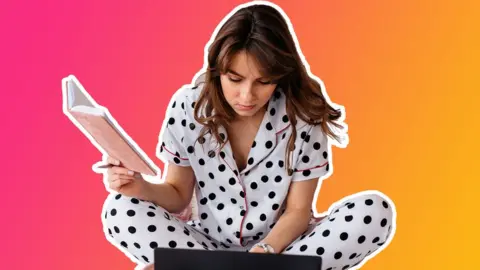 Getty Images A woman in her pyjamas holds a notebook while reading from a laptop, cut out against a pink and orange gradient