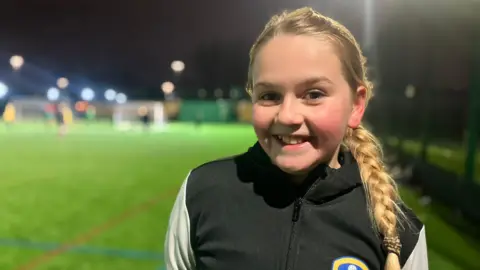 A 11 year old girl with lond blonde hair. She wears a black and white Leeds United suit