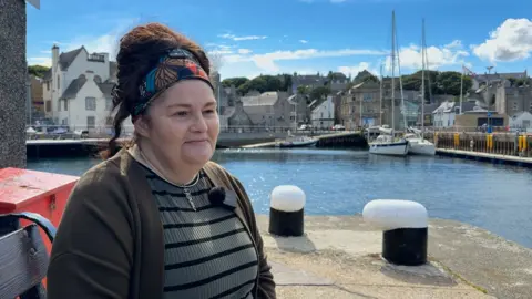 Diane Barnes has dark hair, she is sitting in a harbour area, wearing a green top, necklace and a bandana.