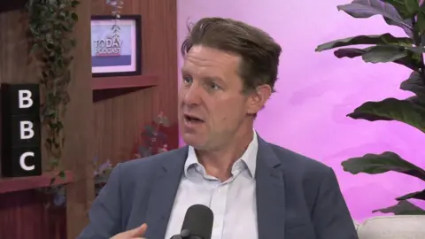 A man, Fraser Nelson, in a BBC studio - he is wearing a grey suit jacket with an unbuttoned white shirt