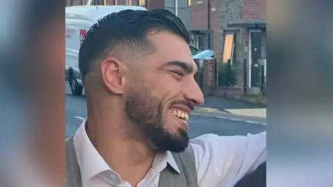 Sadiq Al-lami is seen smiling in a picture taken side-on. He is stood outside wearing a white shirt and grey waistcoat.  