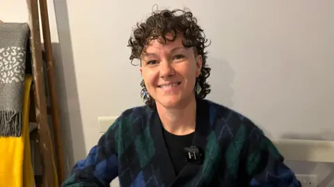 A woman sat down at a table with a clear wall background and hanging blankets to her left. She is wearing a navy tartan cardigan.