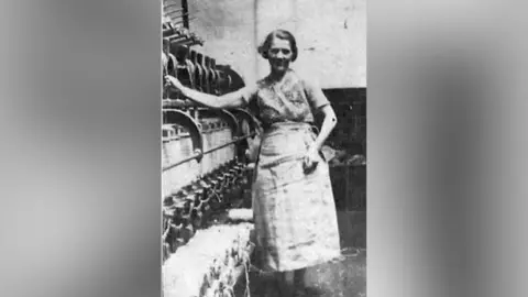 Shirley McMichael A black a white photo of a woman with dark hair. She's standing with one hand on late 1930s machinery and another on her apron, which is tied around her waist over her skirt.