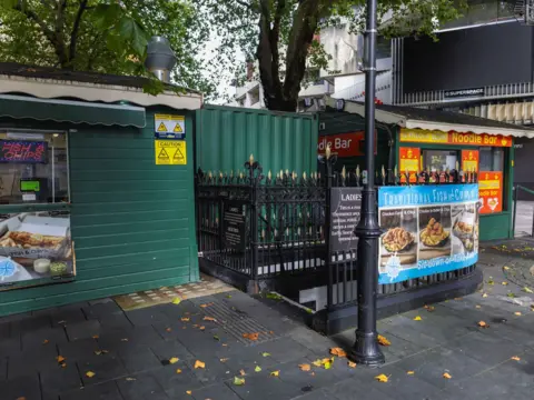 David Hurn The exterior of a subterranean public toilet. Black railings surround concrete steps on a street next to a green hut selling fish and chips and one selling noodles.