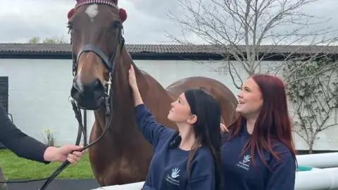 Two girls look up at a brown horse as they stroke its neck. Both girls are wearing blue jumpers with a light blue hand-shape logo. One has black hair, the other dark red hair.