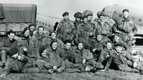 A black and white image of a group of soldiers sitting on the grass in uniform in front of a lorry and an aircraft. 