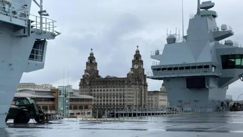 HMS Prince of Wales docked in Liverpool. The ship has a bridge where the time lapse footage of its arrival was filmed from. The deck is empty. Liverpool's Royal Liver building can be seen in the background