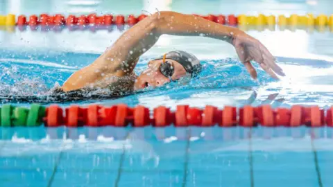 A female swimmer in a black hat and goggles is doing the front crawl.
