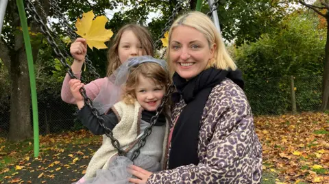 Sam Dixon-French/BBC Katy Irwin has blond, shoulder-length hair. She is wearing a leopard-print coat and is smiling at the camera with her two daughters.