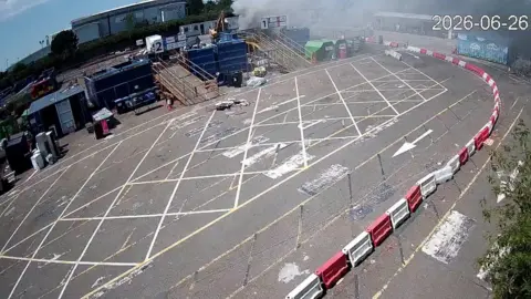 A waste site, with smoke coming from a container, and white and red bollards around it. There are rubbish containers and cabins to the back of the site. 
