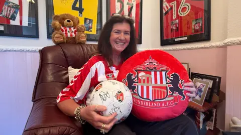 Michelle Barraclough, pictured, has brown hair and is wearing a red and white Sunderland shirt, while holding up a large red Sunderland AFC branded cushion and a white football, which has been signed by various footballers. Behind her are four framed football shirts.