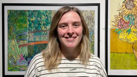 Amelia Riley/BBC News A woman with shoulder-length blonde hair and blue eyes smiles into the camera. She is wearing a black and white striped t-shirt and stands in front of a wall of nature artwork.