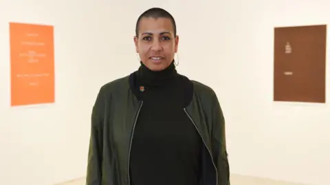 Getty Images/Turner Contemporary Helen Cammock stands looking at the camera in a jacket, in front of an orange and brown poster and white wall. 
