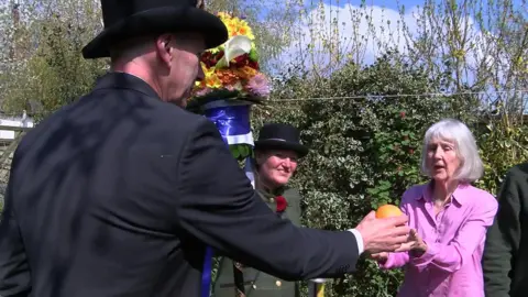 A Tutti man and a Tutti woman visiting a woman and giving her an orange. It is a sunny day. The Tutties are in their traditional attire.
