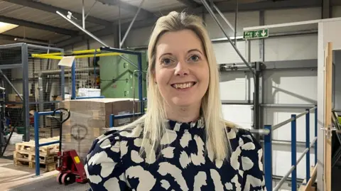 A blonde-haired woman wearing a black and white blouse, standing inside an industrial unit with a pallet truck and railings behind her.