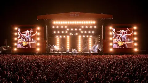 A view of the Radio 2 in the Park festival stage from far back. Thousands of people stand in front of the crowd watching Sting perform at night. The stage is lit up and has two large screens either side.