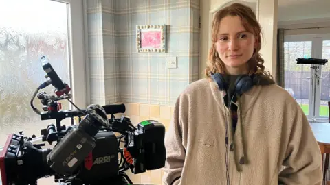 A camera on a tripod is on the left of the photo and Megan stands to the right. She wears a camel coloured fleece and wears a pair of headphones around her neck.