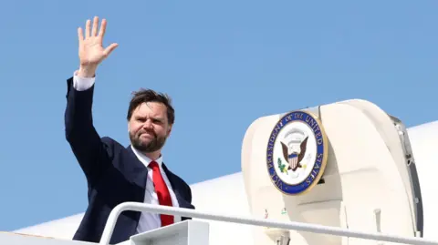 US vice president JD Vance waves before boarding a plane. The US vice president seal features prominently on the plane's door behind him.