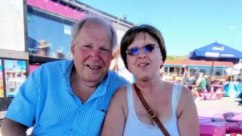 A man with greying hair is wearing a blue and white checked shirt. He is sitting next to a woman with dark blonde hair and a white singlet. She has a handbag strapped across her body and eating a strawberry ice cream. They are sitting at a kiosk at a seafront. 