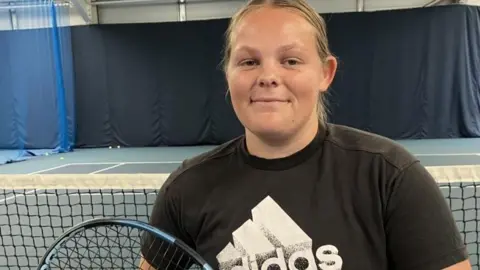 Ruby Bishop at a tennis court. She has blonde, swept-back hair and is wearing a black T-shirt with a large white Adidas logo. She is holding a tennis racket and looking direct at the camera.