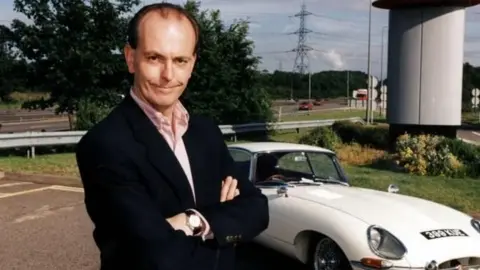 BBC Quentin Willson in 1998 - dressed in a dark blazer, and pink shirt, and standing with his arms crossed  in front of a while vintage Jaguar car  