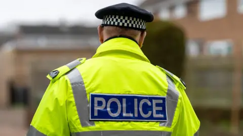A policeman, wearing a yellow hi-vis jacket with the word police on his back. He is also wearing a black and white hate.