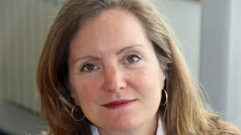 Laura Shoaf has shoulder-length light brown hair. She is wearing small hoop hearings and is slightly smiling towards the camera