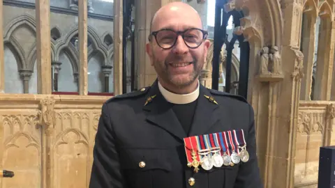 Alan Webber BBC A bearded man stands in front of stonework at Lincoln Cathedral, wearing a blue RAF uniform, medals, a clerical collar and glasses.