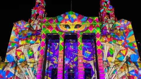 A brightly multi-coloured light installation imposed onto a civic building at night. 