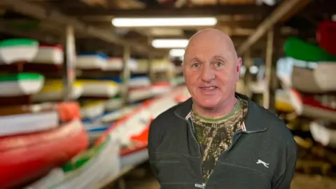 Martin Giles/BBC Craig Block is standing in the boat house and there are multi-coloured boats in the background in rows stacked in the shed. He is wearing a grey jumper with a white logo and a camouflage top underneath. 