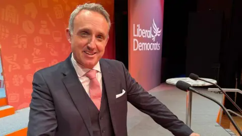 PA Media Alex Cole-Hamilton, who has short grey hair, poses at a podium with orange LibDem branding in the background 