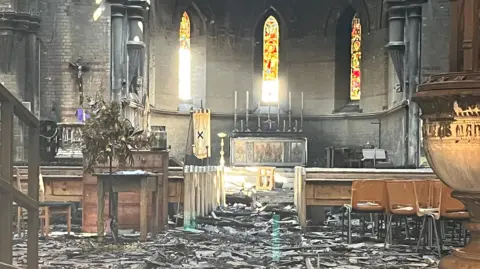 Interior of church damaged by fire. Three stained glass windows are in the background and fire damaged items are on the ground.