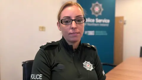 A picture of PSNI Chief Superintendent Amanda Ford.
She is wearing the green PSNI uniform which has a quarter zip and the PSNI emblem on the front.
She has blonde hair tied back in a ponytail and is wearing black framed glasses.