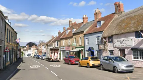 A street view image of Wincanton High Street. The road is lined with cars, and flanked by thatched houses and shops.