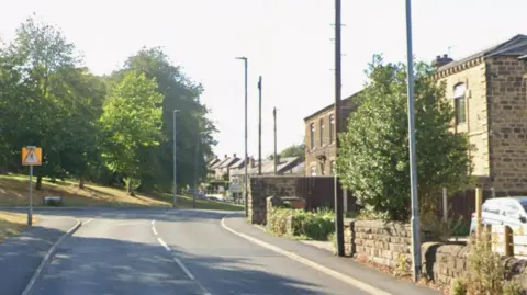 Google A single carriageway road with buildings on the right side and a pavement and grass bank with trees on the left side.A junction turn off left ahead.