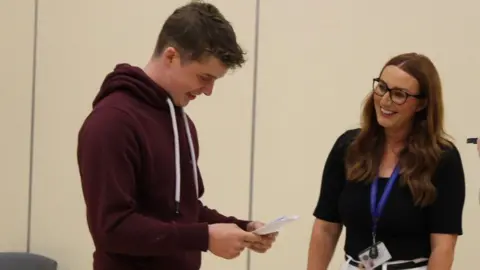 Lecale Trinity Grammar School A young man in a maroon hooded top is looking at a sheet of paper that contains his exam results. He is smiling. A woman with dark hair and glasses in a dark top is also smiling.