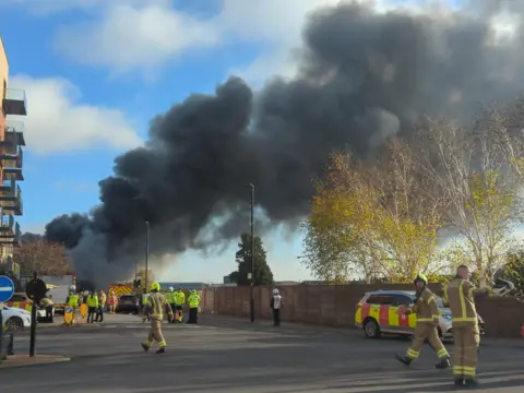 Large plume of black smoke emerges from a building (left)
In the foreground are firefighters in uniform including yellow helmets.