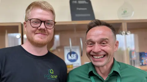 Two men stand side by side, both smiling. On the left, Aran has ginger hair and a ginger beard. He is also wearing a pair of rounded burgundy glasses. Grant, an older man, has brown hair and wears a green shirt. Behind them, a blurred view of a cabinet with a display of company awards.