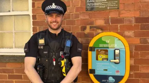 Devon and Cornwall Police PC Ross Buckler stands next to a defibrillator installed outside Sidmouth police station. He is wearing police uniform.