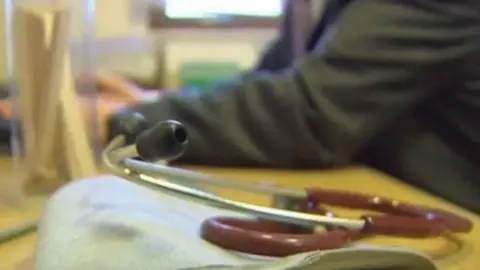 BBC A stethoscope on a GP's desk with the doctor sitting at a yellow desk.